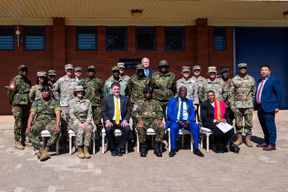 A formal group photograph taken outdoors in front of a brick building, featuring a mixed group of military personnel in various camouflage uniforms and civilians in business attire. The group appears to represent multiple nationalities and military organizations, suggesting an international military cooperation or partnership event. Participants are arranged in two rows, with senior figures seated in the front row.