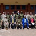 A formal group photograph taken outdoors in front of a brick building, featuring a mixed group of military personnel in various camouflage uniforms and civilians in business attire. The group appears to represent multiple nationalities and military organizations, suggesting an international military cooperation or partnership event. Participants are arranged in two rows, with senior figures seated in the front row.