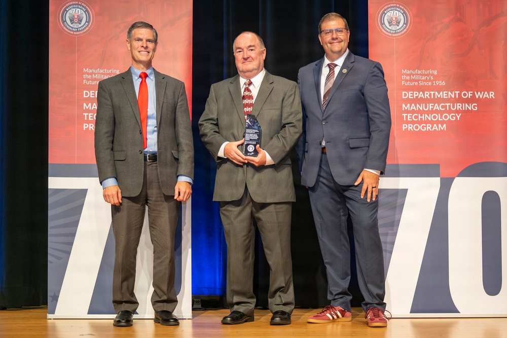 Three men in business attire are posed on a stage at what appears to be an awards ceremony for the Department of War Manufacturing Technology Program. The central figure is holding a crystal or glass award trophy. The backdrop features red banners with program branding and the number '70,' suggesting a 70th anniversary celebration.