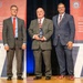 Three men in business attire are posed on a stage at what appears to be an awards ceremony for the Department of War Manufacturing Technology Program. The central figure is holding a crystal or glass award trophy. The backdrop features red banners with program branding and the number '70,' suggesting a 70th anniversary celebration.