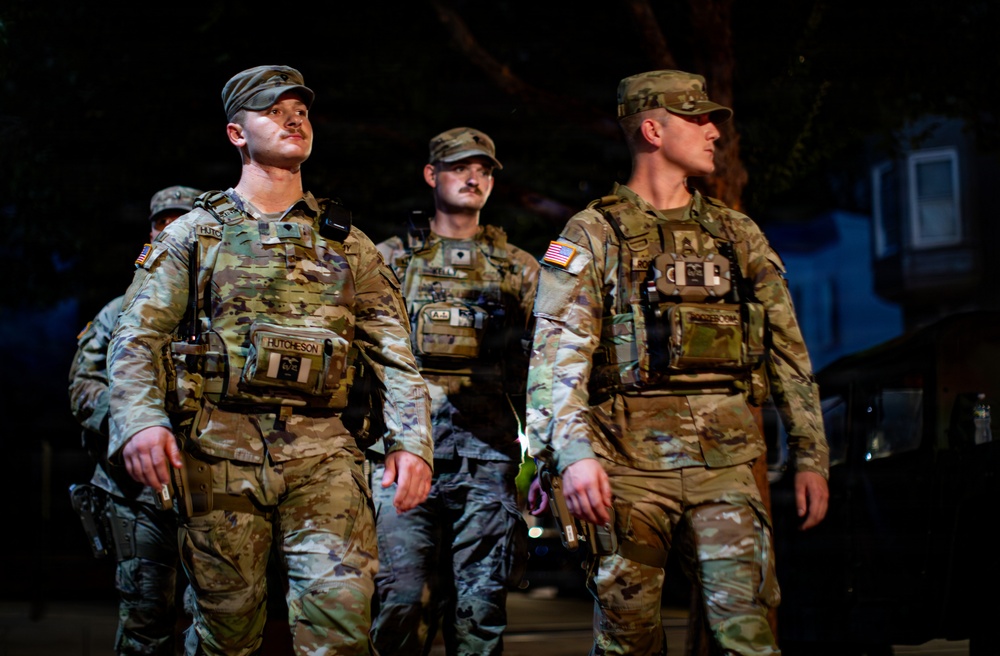 A group of military personnel in full combat gear are photographed at night in what appears to be an urban setting. The soldiers are wearing MultiCam/OCP camouflage uniforms with tactical vests, patrol caps, and name tapes visible. They appear to be on patrol or standing in formation, looking alert.