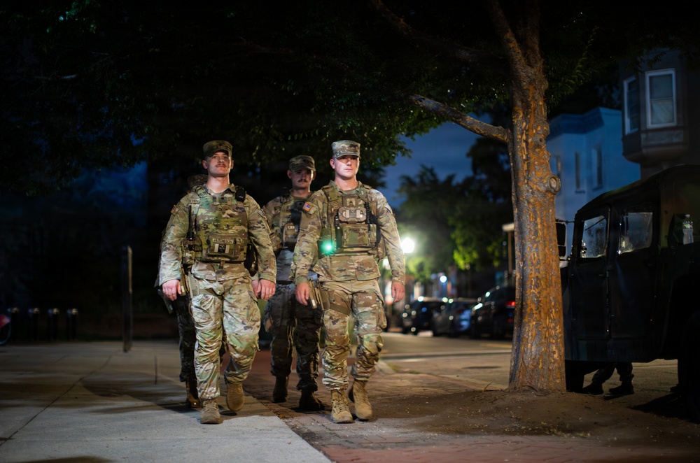 Three military personnel in camouflage uniforms and tactical vests are walking along a sidewalk at night in what appears to be an urban residential area. They are equipped with body armor and appear to be conducting a patrol. A military vehicle, likely a Humvee, is partially visible on the right side of the frame.