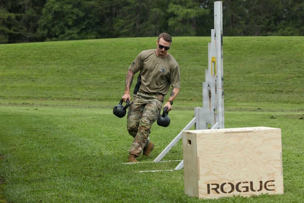 A military service member in OCP (Operational Camouflage Pattern) uniform is performing a fitness challenge on a grass field, carrying two kettlebells in a farmer's carry exercise. The individual is navigating what appears to be a marked course with a Rogue-branded plywood jump box and a metal rack structure visible nearby. The setting suggests a military physical fitness competition or training event conducted outdoors.