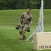 A military service member in OCP (Operational Camouflage Pattern) uniform is performing a fitness challenge on a grass field, carrying two kettlebells in a farmer's carry exercise. The individual is navigating what appears to be a marked course with a Rogue-branded plywood jump box and a metal rack structure visible nearby. The setting suggests a military physical fitness competition or training event conducted outdoors.