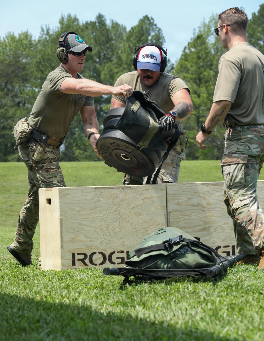 Three military personnel in OCP/multicam uniforms are engaged in a physical fitness or competition event outdoors. They are lifting or maneuvering a heavy object, likely a weighted implement, over a large wooden plywood box branded with 'ROGUE'. A military-style bag or ruck is visible on the grass in the foreground.
