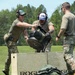 Three military personnel in OCP/multicam uniforms are engaged in a physical fitness or competition event outdoors. They are lifting or maneuvering a heavy object, likely a weighted implement, over a large wooden plywood box branded with 'ROGUE'. A military-style bag or ruck is visible on the grass in the foreground.