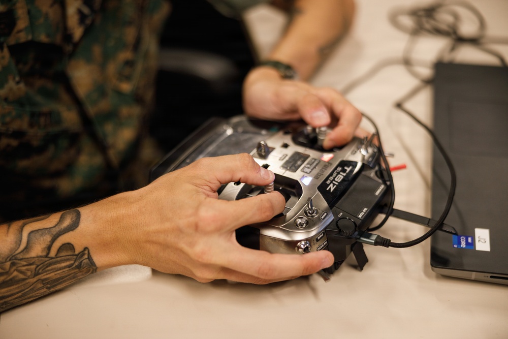 Two individuals are handling a Futaba T16IZ Super RC radio transmitter controller on a white table surface. One person is wearing military camouflage (MARPAT pattern) uniform, and the other has a visible tattoo on their forearm. The controller is connected via cables, possibly to a laptop visible on the right edge of the frame.