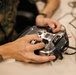 Two individuals are handling a Futaba T16IZ Super RC radio transmitter controller on a white table surface. One person is wearing military camouflage (MARPAT pattern) uniform, and the other has a visible tattoo on their forearm. The controller is connected via cables, possibly to a laptop visible on the right edge of the frame.
