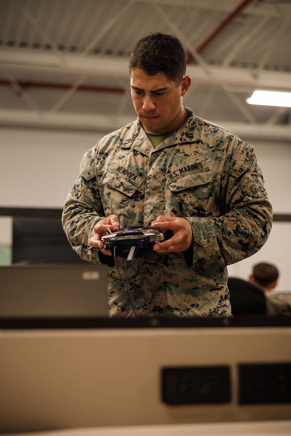 A U.S. Marine identified by name tape as 'Mendoza' stands in what appears to be an indoor training or operations facility, holding a remote control device. He is wearing the Marine Corps MARPAT digital camouflage uniform with 'U.S. MARINES' tape visible on his chest. He appears focused on the controller in his hands, suggesting drone or unmanned system operation training.