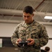 A U.S. Marine identified by name tape as 'Mendoza' stands in what appears to be an indoor training or operations facility, holding a remote control device. He is wearing the Marine Corps MARPAT digital camouflage uniform with 'U.S. MARINES' tape visible on his chest. He appears focused on the controller in his hands, suggesting drone or unmanned system operation training.