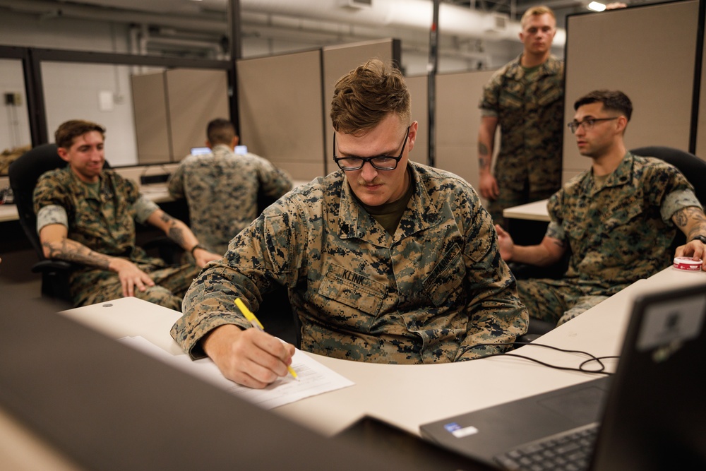 A group of U.S. Marines in MARPAT camouflage uniforms are gathered in what appears to be an office or operations center environment. The central figure, identified by his name tape as 'KLINK,' is focused on writing on a document at a desk while wearing glasses. Several other Marines are visible in the background, some seated and some standing, in a cubicle-style workspace.