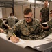 A group of U.S. Marines in MARPAT camouflage uniforms are gathered in what appears to be an office or operations center environment. The central figure, identified by his name tape as 'KLINK,' is focused on writing on a document at a desk while wearing glasses. Several other Marines are visible in the background, some seated and some standing, in a cubicle-style workspace.