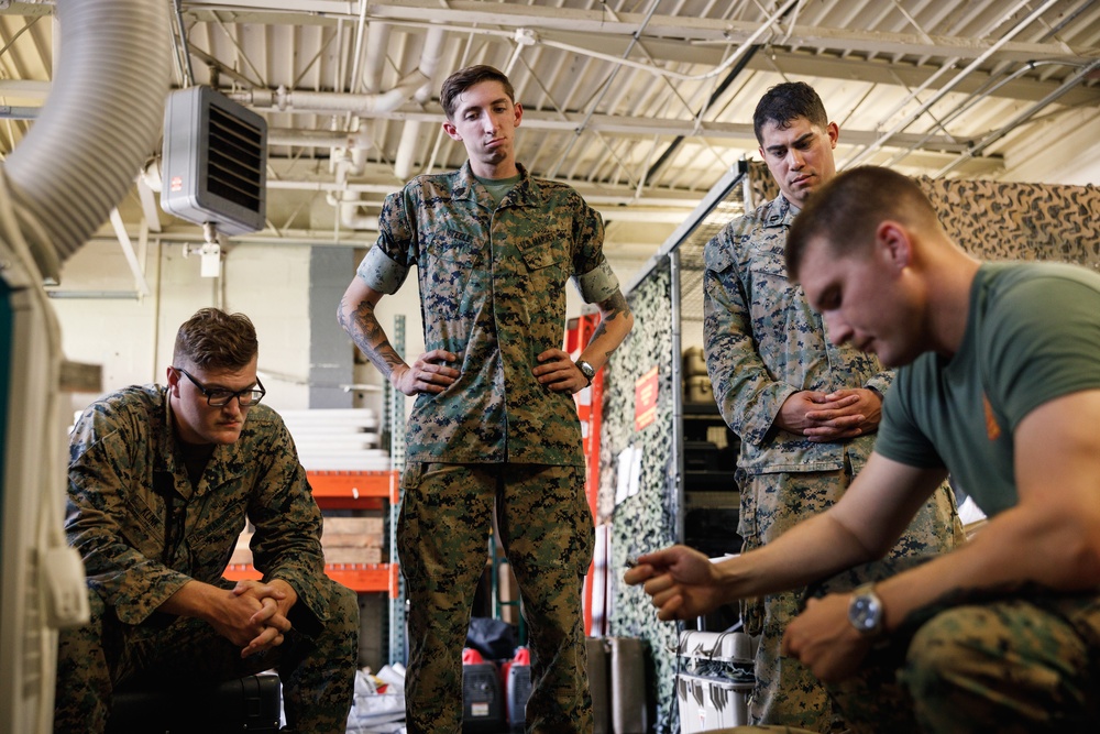 Four military personnel in U.S. Marine Corps MARPAT camouflage uniforms are gathered in what appears to be a maintenance bay or warehouse facility. One individual is actively working on equipment while the others observe, suggesting a training or maintenance demonstration scenario. The setting includes industrial shelving, camouflage netting, and HVAC equipment overhead.