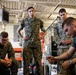 Four military personnel in U.S. Marine Corps MARPAT camouflage uniforms are gathered in what appears to be a maintenance bay or warehouse facility. One individual is actively working on equipment while the others observe, suggesting a training or maintenance demonstration scenario. The setting includes industrial shelving, camouflage netting, and HVAC equipment overhead.