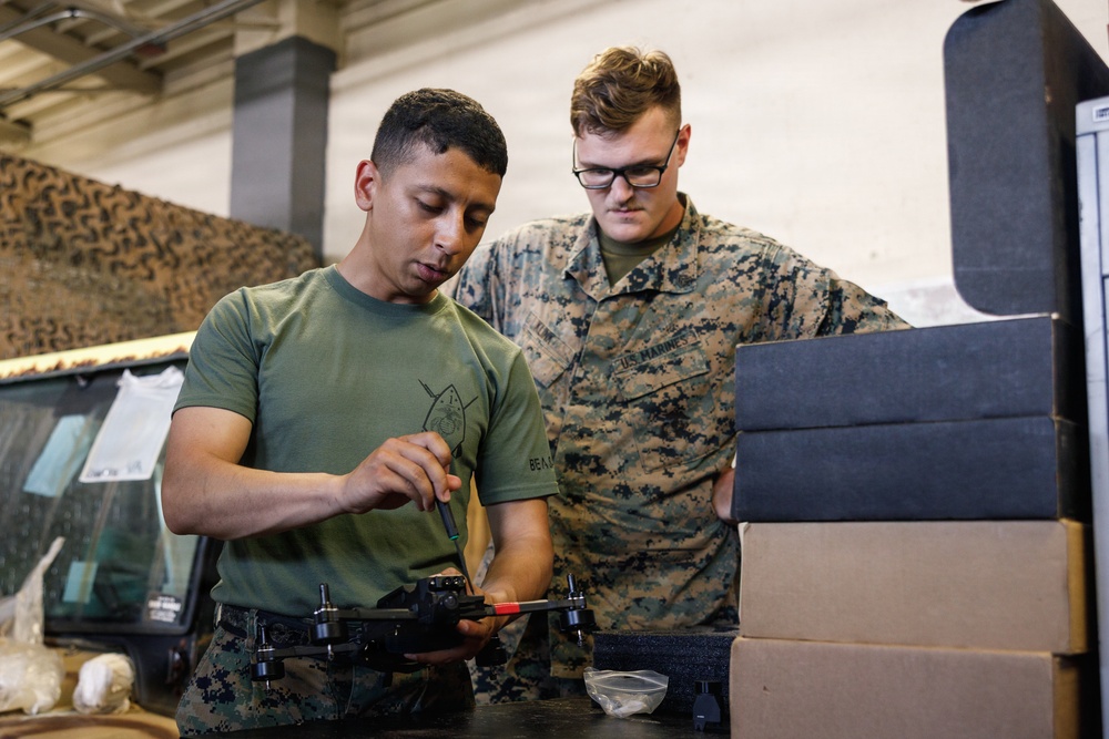 Two U.S. Marines are working together on a small quadcopter drone at a workbench inside what appears to be a military facility or hangar. One Marine in a green t-shirt is handling the drone with a screwdriver, while the other in full MARPAT camouflage uniform observes. Cardboard and foam packaging boxes are stacked nearby, suggesting the equipment may be newly issued or being prepared for use.