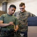 Two U.S. Marines are working together on a small quadcopter drone at a workbench inside what appears to be a military facility or hangar. One Marine in a green t-shirt is handling the drone with a screwdriver, while the other in full MARPAT camouflage uniform observes. Cardboard and foam packaging boxes are stacked nearby, suggesting the equipment may be newly issued or being prepared for use.