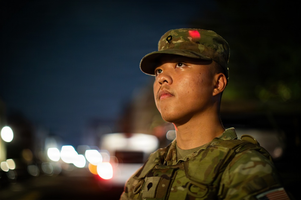 A young soldier in U.S. Army Operational Camouflage Pattern (OCP) uniform is photographed at night in an urban setting. The subject is gazing upward and to the left with a composed expression, illuminated by ambient street lighting. The background shows bokeh street lights suggesting a busy urban environment.