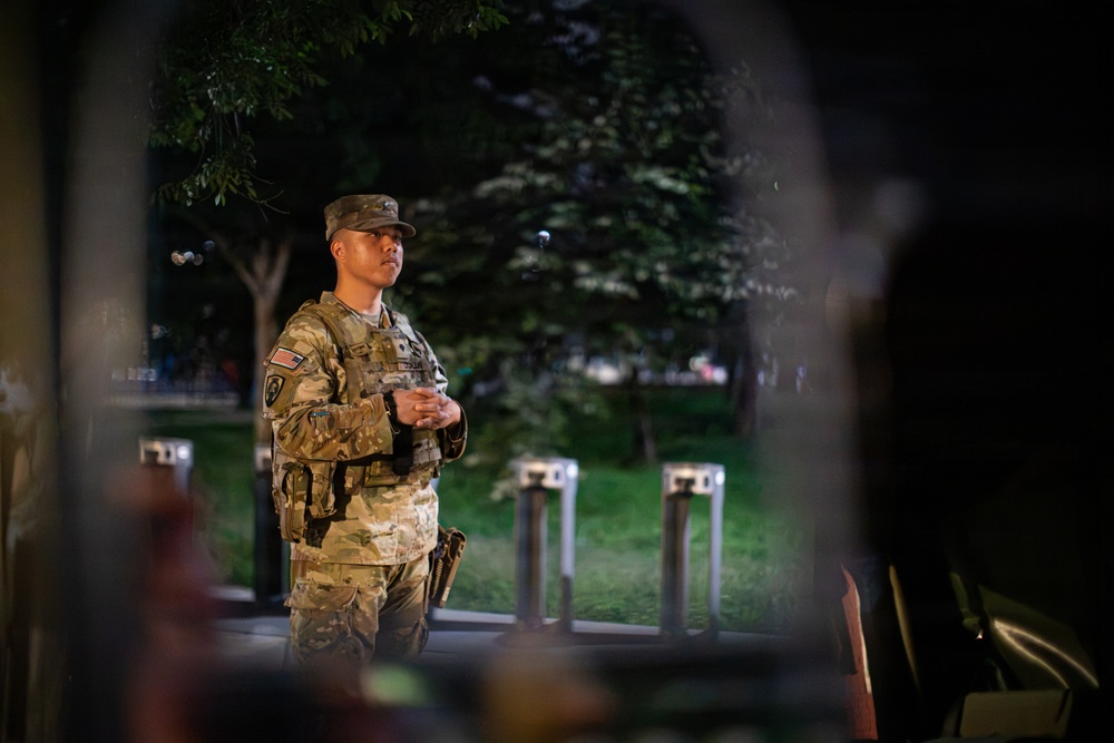 A U.S. Army soldier in full multicam combat uniform and tactical vest stands at attention during nighttime hours, photographed through what appears to be a gap in a fence or barrier. The soldier appears to be on guard duty at what looks like a secured perimeter, with bollards and a green park area visible in the background. The image is taken with a shallow depth of field, creating a framed, documentary-style composition.