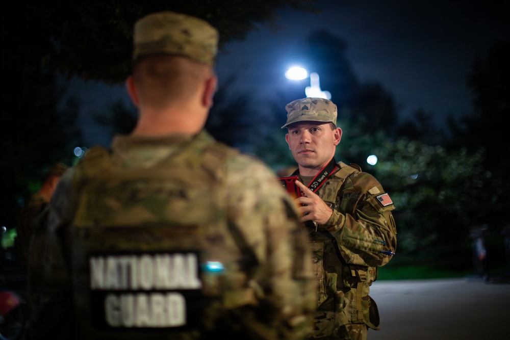 Two National Guard soldiers are engaged in a nighttime briefing or conversation in an outdoor setting. The soldier facing the camera holds a red mobile device and wears Sergeant rank insignia and a US flag patch on his right sleeve. The scene appears to be an operational deployment or security mission.