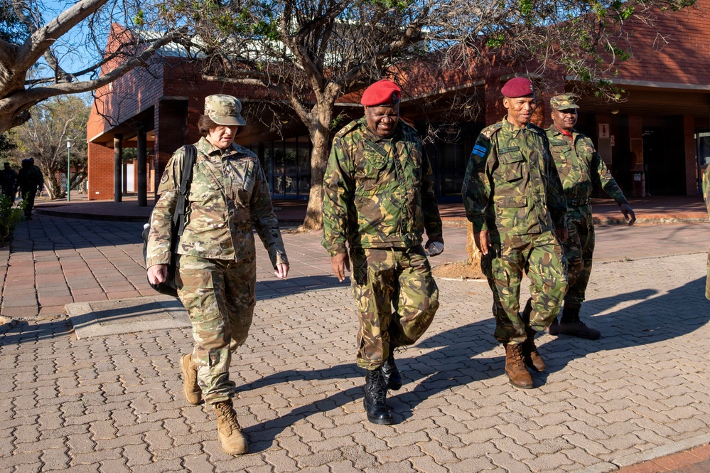 Four military personnel are walking together on a brick-paved outdoor area near a red-brick building. One individual on the left wears US Army MultiCam (OCP) uniform with a patrol cap, while the other three wear green camouflage uniforms with red berets, suggesting a multinational military engagement or liaison visit. The setting appears to be a military installation or institutional campus in a warm, arid environment.