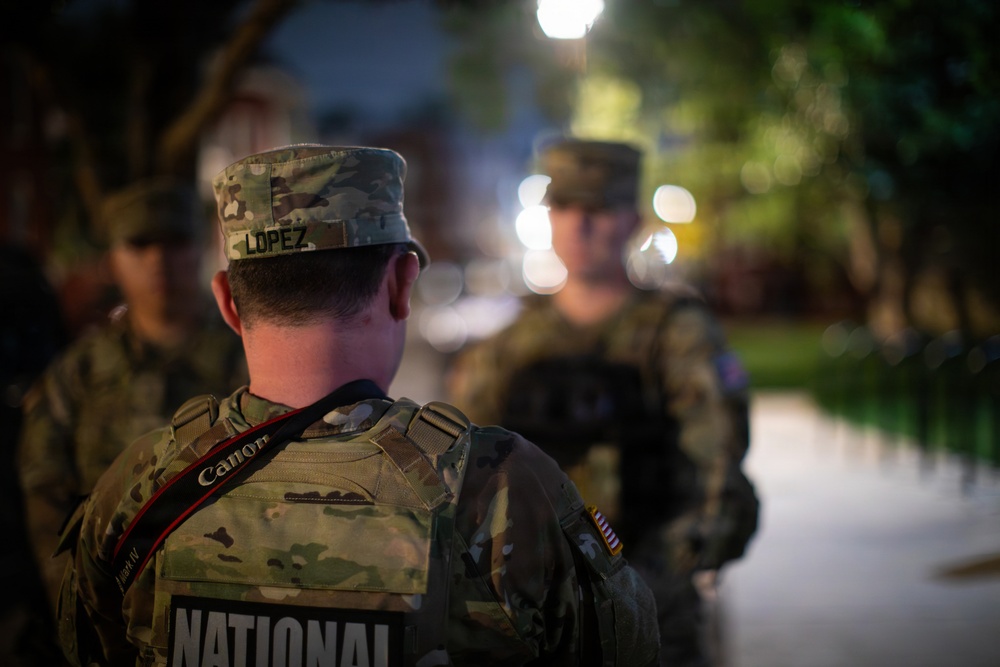 The image shows military personnel in camouflage uniforms at night, with the foreground soldier identified by the name tape 'LOPEZ' on their cap. The word 'NATIONAL' is visible on the back of the foreground soldier's vest, indicating National Guard affiliation. A Canon camera strap is visible across the soldier's back, suggesting a documentation or media role.