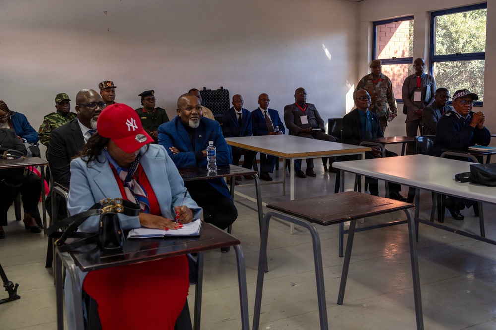 A formal meeting or briefing is taking place in a classroom-style room with multiple attendees seated at desks and some standing along the walls. The group includes individuals in both civilian attire and military/police uniforms, suggesting an official or governmental gathering. Participants appear to be listening to a presentation or speaker located outside the frame.