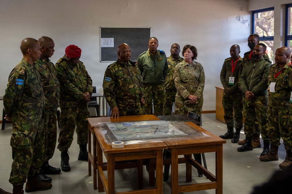 A group of military personnel from multiple nations are gathered around a table with a map during what appears to be a briefing or planning session in a classroom or operations room. One individual in camouflage uniform appears to be speaking or presenting to the group. The presence of different uniform styles and flag patches suggests a multinational military exercise or coordination meeting.