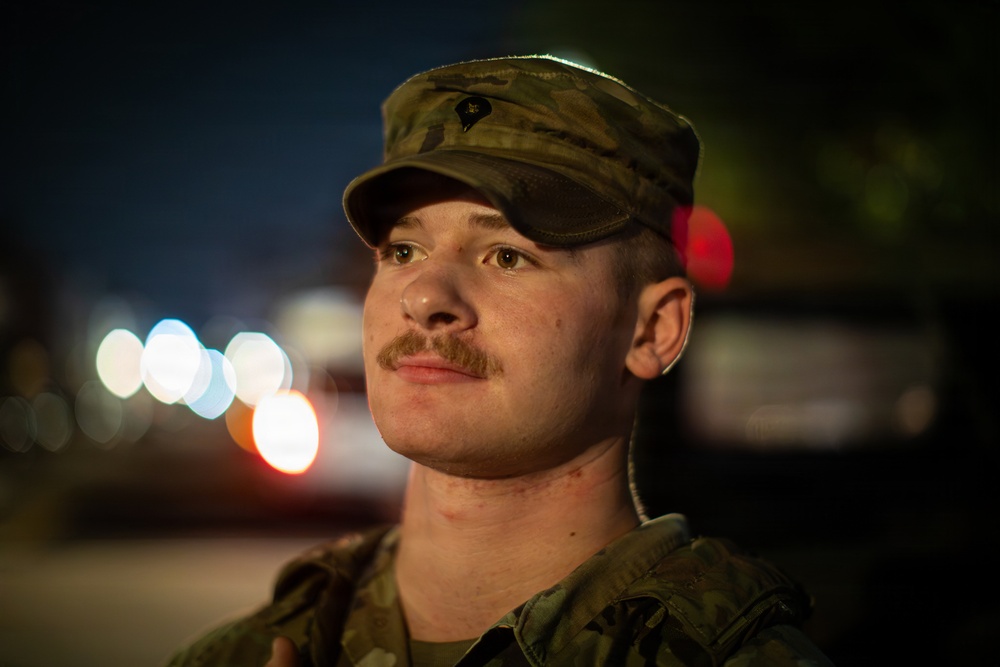 A young male soldier wearing a camouflage uniform and patrol cap is photographed at night in a close-up portrait. He has a mustache and is looking upward and to the side. The background shows bokeh lights consistent with a nighttime outdoor urban or base environment.