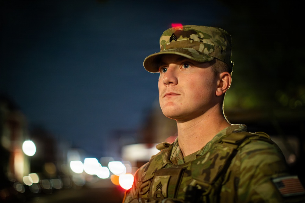 A young male soldier in U.S. Army uniform is photographed at night in what appears to be an urban setting. He is wearing MultiCam (OCP) camouflage pattern uniform with a patrol cap bearing a Staff Sergeant rank insignia and a tactical vest. The image is a close-up portrait with a shallow depth of field, with bokeh street lights visible in the background.