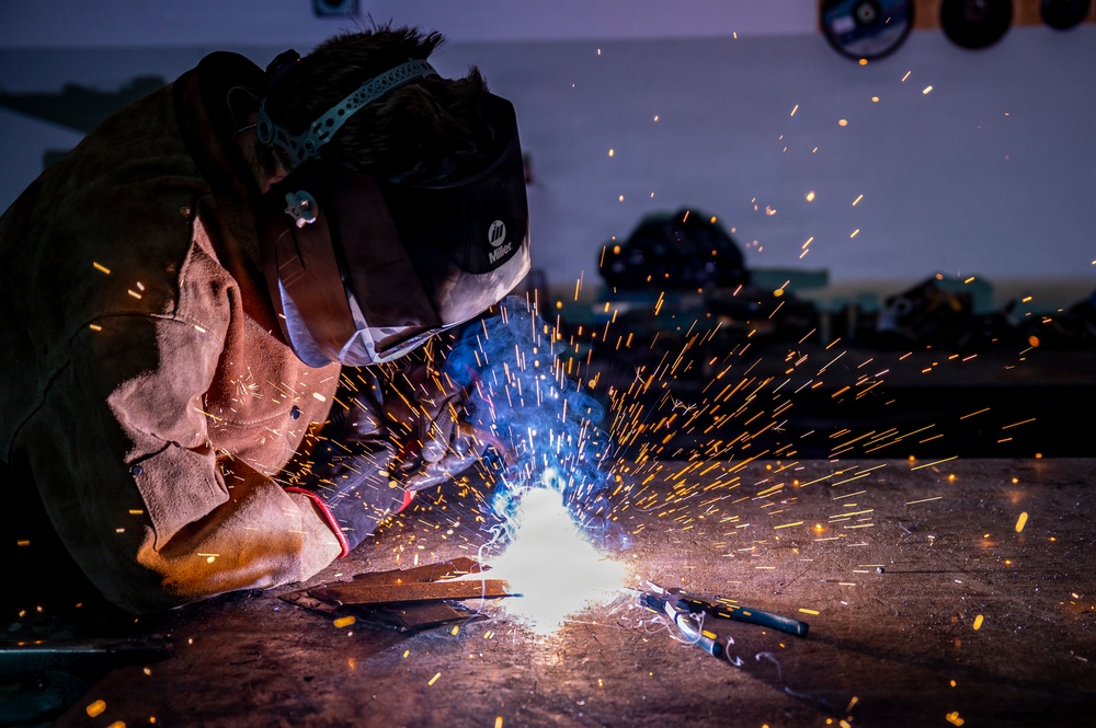 A welder is actively welding metal on a flat surface in what appears to be an industrial workshop or fabrication facility. The image captures a dramatic spray of orange and white sparks emanating from the welding arc, with a bright blue-white light at the point of contact. The welder is wearing full protective gear including a Miller-brand welding helmet and a leather welding jacket.