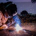 A welder is actively welding metal on a flat surface in what appears to be an industrial workshop or fabrication facility. The image captures a dramatic spray of orange and white sparks emanating from the welding arc, with a bright blue-white light at the point of contact. The welder is wearing full protective gear including a Miller-brand welding helmet and a leather welding jacket.