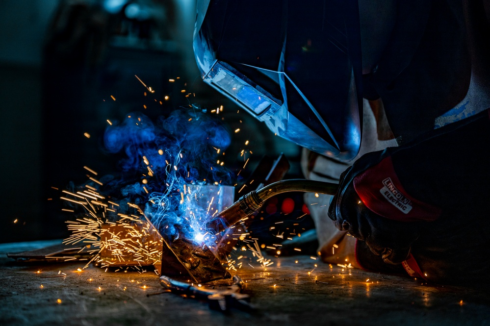 A welder is actively welding metal pieces on a workshop floor, producing a bright arc with flying sparks and blue smoke. The worker is equipped with professional safety gear including a welding helmet and branded gloves. The dramatic low-angle composition captures the intensity of the welding process in a dark industrial environment.