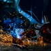 A welder is actively welding metal pieces on a workshop floor, producing a bright arc with flying sparks and blue smoke. The worker is equipped with professional safety gear including a welding helmet and branded gloves. The dramatic low-angle composition captures the intensity of the welding process in a dark industrial environment.