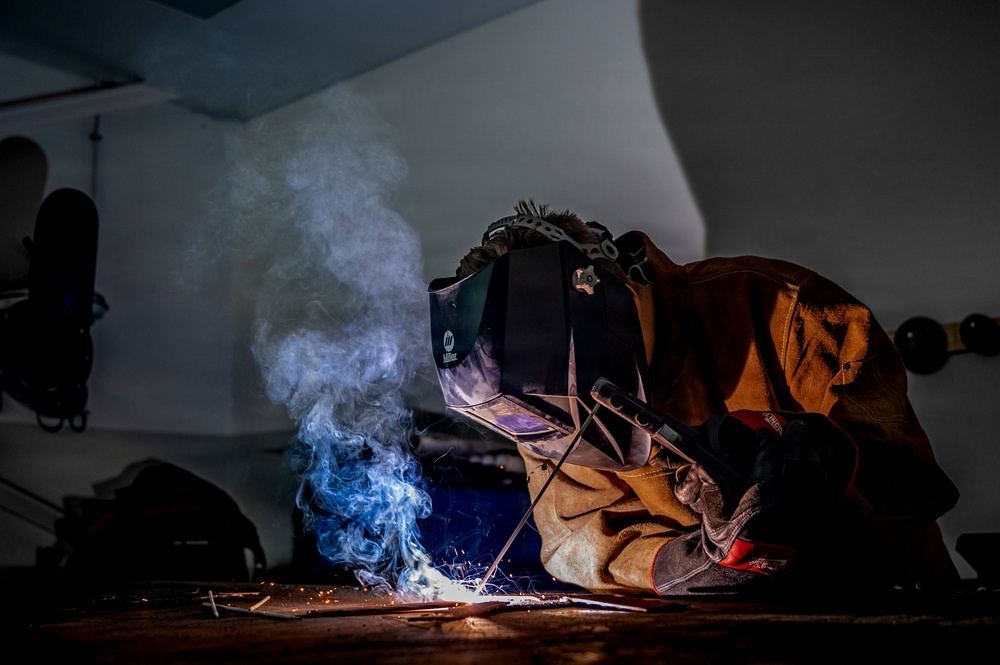 A welder is actively welding metal on a workbench in what appears to be a workshop or garage setting. The welder is wearing full protective gear including a Miller auto-darkening welding helmet, leather welding jacket, and leather gloves. Bright welding sparks and blue-tinted smoke are visible, dramatically illuminating the otherwise dark workspace.