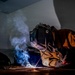 A welder is actively welding metal on a workbench in what appears to be a workshop or garage setting. The welder is wearing full protective gear including a Miller auto-darkening welding helmet, leather welding jacket, and leather gloves. Bright welding sparks and blue-tinted smoke are visible, dramatically illuminating the otherwise dark workspace.