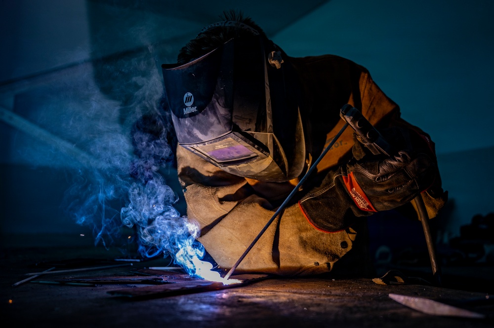 A welder is actively performing a welding operation on a metal surface in a dark industrial environment. The worker is equipped with a Miller brand auto-darkening welding helmet, leather welding jacket, and protective gloves. The welding arc produces a vivid blue-white flame with visible smoke and sparks, dramatically illuminating the scene.
