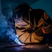 A welder is actively performing a welding operation on a metal surface in a dark industrial environment. The worker is equipped with a Miller brand auto-darkening welding helmet, leather welding jacket, and protective gloves. The welding arc produces a vivid blue-white flame with visible smoke and sparks, dramatically illuminating the scene.