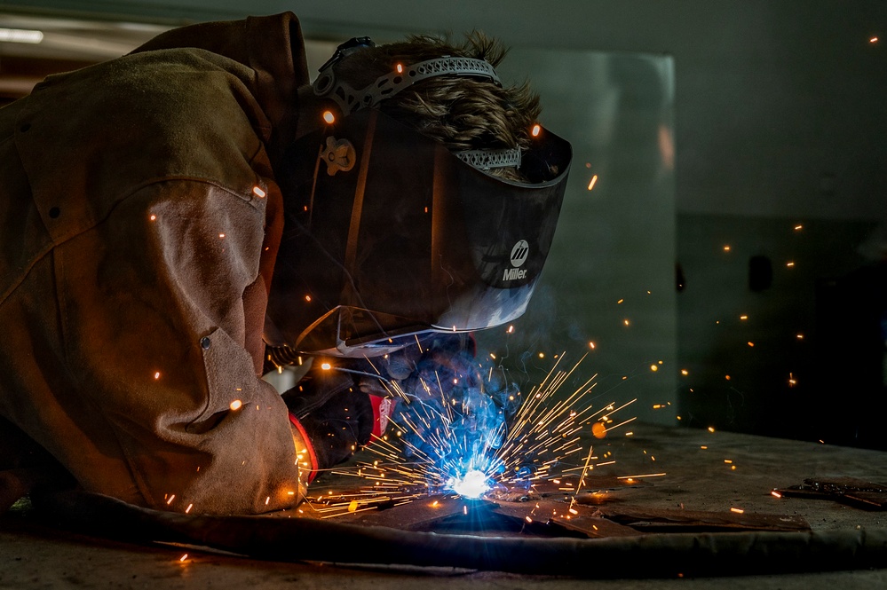A welder is actively welding metal on a work surface in what appears to be an industrial or workshop setting. The welder is wearing a Miller brand auto-darkening welding helmet and a leather welding jacket for protection. Bright blue-white arc light and numerous orange sparks are visible, indicating active metal fusion in progress.