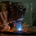 A welder is actively welding metal on a work surface in what appears to be an industrial or workshop setting. The welder is wearing a Miller brand auto-darkening welding helmet and a leather welding jacket for protection. Bright blue-white arc light and numerous orange sparks are visible, indicating active metal fusion in progress.