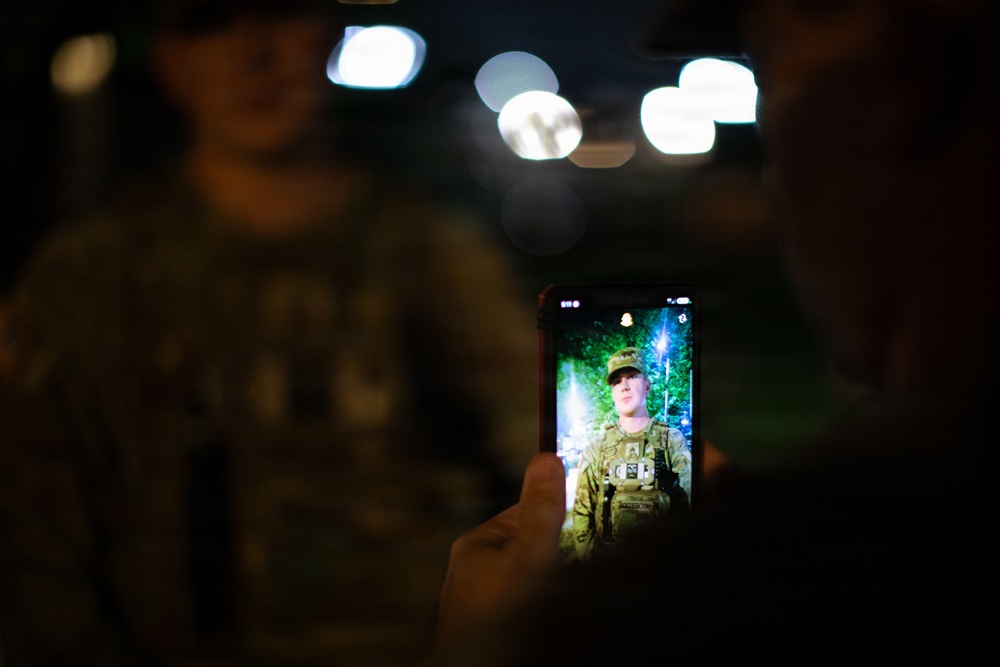 A smartphone is being held up in a dark environment, displaying a photo of a soldier in military camouflage uniform and tactical vest. The background of the main scene is dark with bokeh light effects, while a blurred figure in military attire is visible in the foreground. The phone screen serves as the primary light source and focal point of the image.