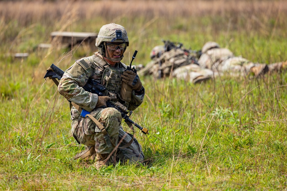 A U.S. Army soldier identified by name tape as 'SMITH' is kneeling in a grassy field while communicating on a handheld radio. The soldier is in full combat gear including OCP (Operational Camouflage Pattern) uniform, tactical vest, helmet, and protective eyewear, and is holding an M4-style rifle. In the background, additional soldiers are visible in prone positions, suggesting a field training exercise.