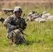A U.S. Army soldier identified by name tape as 'SMITH' is kneeling in a grassy field while communicating on a handheld radio. The soldier is in full combat gear including OCP (Operational Camouflage Pattern) uniform, tactical vest, helmet, and protective eyewear, and is holding an M4-style rifle. In the background, additional soldiers are visible in prone positions, suggesting a field training exercise.