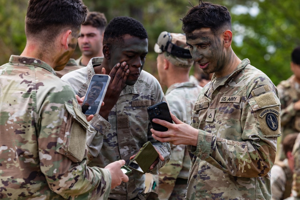 A group of U.S. Army soldiers in camouflage uniforms are applying or checking face paint/camouflage makeup. Two soldiers in the foreground are using smartphones as mirrors to inspect their camouflage face paint application. The setting appears to be an outdoor training environment with trees visible in the background.