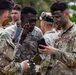 A group of U.S. Army soldiers in camouflage uniforms are applying or checking face paint/camouflage makeup. Two soldiers in the foreground are using smartphones as mirrors to inspect their camouflage face paint application. The setting appears to be an outdoor training environment with trees visible in the background.