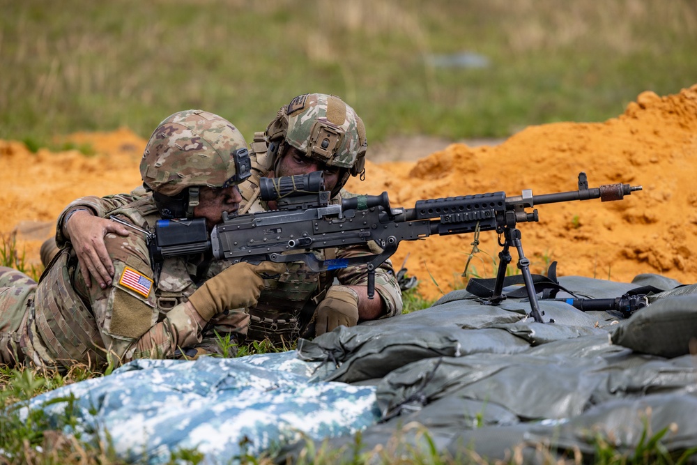 Two U.S. Army soldiers in full combat gear are positioned prone behind sandbags at what appears to be a live-fire training range. One soldier is aiming an M240 machine gun mounted on a bipod while the other provides close instruction or guidance. The presence of orange/red disturbed earth in the background and sandbag fortifications suggests a controlled training environment.