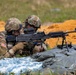 Two U.S. Army soldiers in full combat gear are positioned prone behind sandbags at what appears to be a live-fire training range. One soldier is aiming an M240 machine gun mounted on a bipod while the other provides close instruction or guidance. The presence of orange/red disturbed earth in the background and sandbag fortifications suggests a controlled training environment.