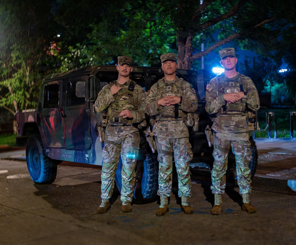 Three military personnel in MultiCam/OCP camouflage uniforms are posed in front of a military HMMWV (Humvee) at night. They are equipped with tactical gear including plate carriers, holstered sidearms, and radio equipment. The scene is illuminated by artificial lighting against a backdrop of trees.