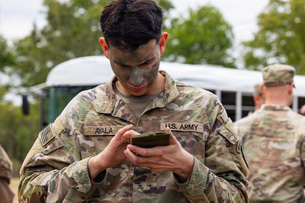 A U.S. Army soldier named Ayala, wearing OCP (Operational Camouflage Pattern) uniform with face camouflage paint applied, is looking down at a small green military-style notebook or device held in both hands. The soldier is standing outdoors near a white bus with trees visible in the background. A second soldier in OCP uniform is partially visible in the background to the right.