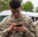 A U.S. Army soldier named Ayala, wearing OCP (Operational Camouflage Pattern) uniform with face camouflage paint applied, is looking down at a small green military-style notebook or device held in both hands. The soldier is standing outdoors near a white bus with trees visible in the background. A second soldier in OCP uniform is partially visible in the background to the right.
