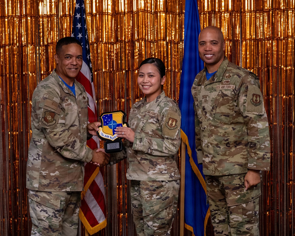 Three U.S. Air Force personnel in OCP (Operational Camouflage Pattern) uniforms pose for a formal award photo. The central figure is receiving a shield-shaped plaque bearing what appears to be the '7th Air Base Wing' emblem from the individual on the left, identified by nametape as 'HARRIS'. The ceremony takes place in front of an American flag and a blue Air Force flag against a gold tinsel backdrop.